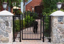 Custom iron walk gate with integrated initial and puppy pickets mounted between stone columns (Style #15 - Puppy Picket Finials)